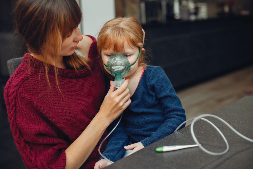Mother helps daughter with asthma therapy, wearing a mask for inhalation