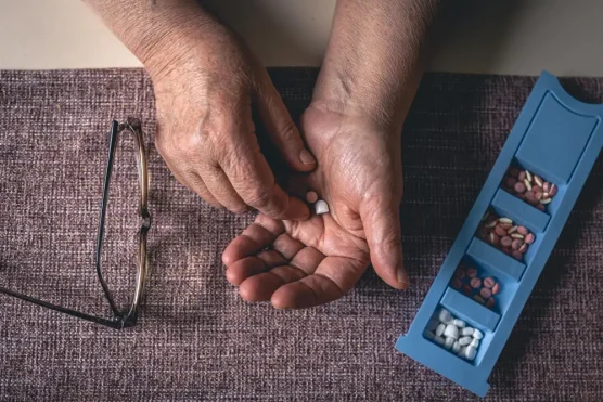 Senior person organizing daily medications with pill organizer and glasses on table, representing the need to buy Cialis from Canada for affordable treatment.