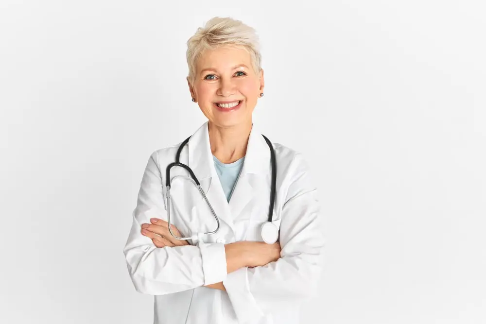 A smiling female doctor with short, light-colored hair, wearing a white lab coat and a stethoscope around her neck, stands with her arms crossed, representing a Canadian pharmacy that fills U.S. prescriptions.