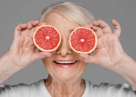 Smiling senior woman playfully holding grapefruit halves over her eyes, symbolizing the importance of understanding Eliquis food interactions, particularly with grapefruit.