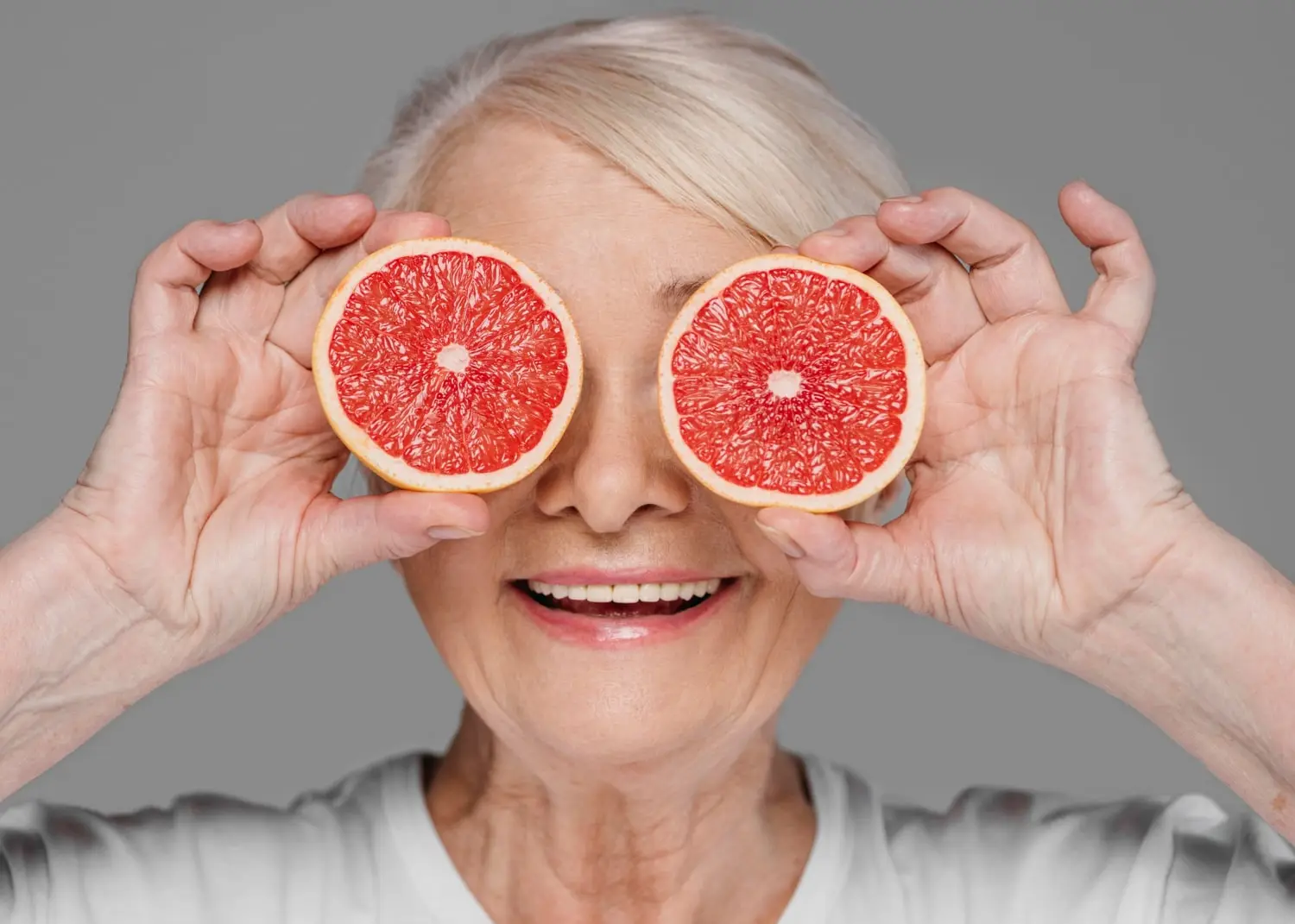 Smiling senior woman playfully holding grapefruit halves over her eyes, symbolizing the importance of understanding Eliquis food interactions, particularly with grapefruit.