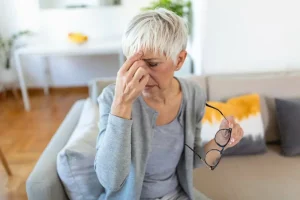 Senior woman holding her head in discomfort while sitting on a couch, symbolizing migraine pain and concern about Nurtec ODT price.