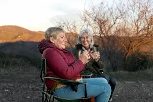 Two cheerful senior women laughing and holding mugs while sitting in folding chairs outdoors, representing healthy aging and discussions about generic Eliquis for stroke prevention.