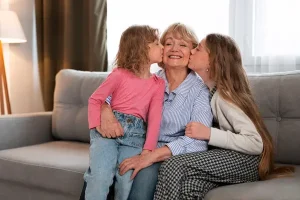 A smiling senior grandmother sitting on a couch receiving kisses on the cheek from her two granddaughters, used to illustrate health safety and how long does Eliquis stay in your system.