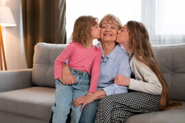 A smiling senior grandmother sitting on a couch receiving kisses on the cheek from her two granddaughters, used to illustrate health safety and how long does Eliquis stay in your system.
